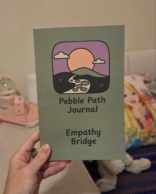 A hand holds a square journal cover titled 'Pebble Path Journal' with the subtitle 'Empathy Bridge.' The cover is muted sage green with a central illustrated scene showing a purple sky, peach sun, dark hills, and a white rabbit.