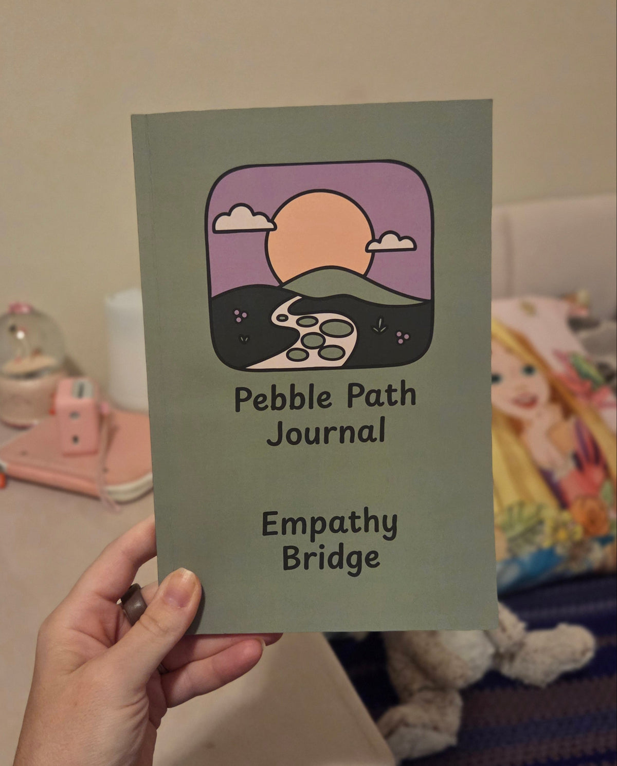 A hand holds a square journal cover titled 'Pebble Path Journal' with the subtitle 'Empathy Bridge.' The cover is muted sage green with a central illustrated scene showing a purple sky, peach sun, dark hills, and a white rabbit.